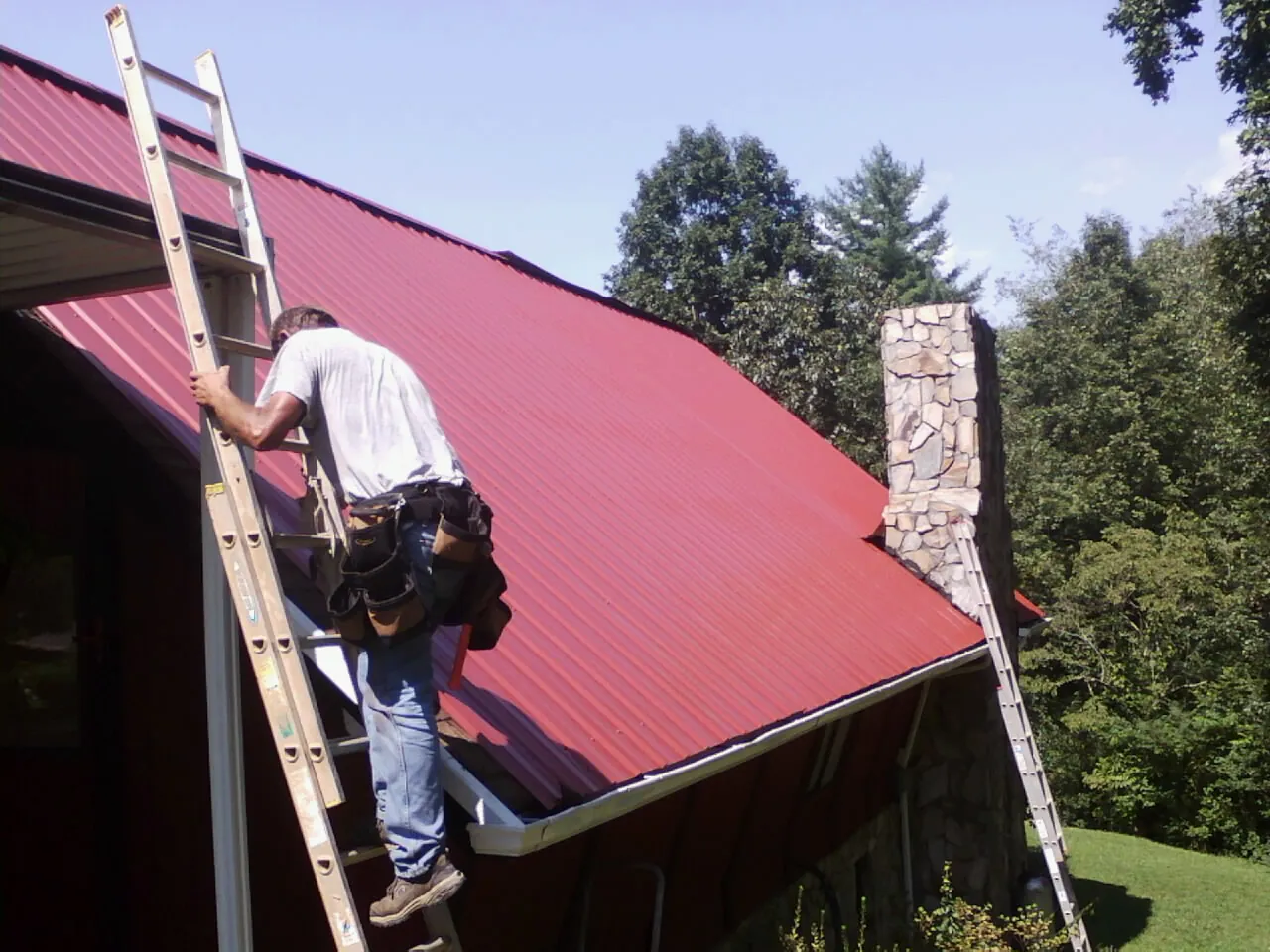 Roofer on a ladder beside a red metal roof and stone chimney — Soffit & Fascia Repair by Roofing Brothers in Montour